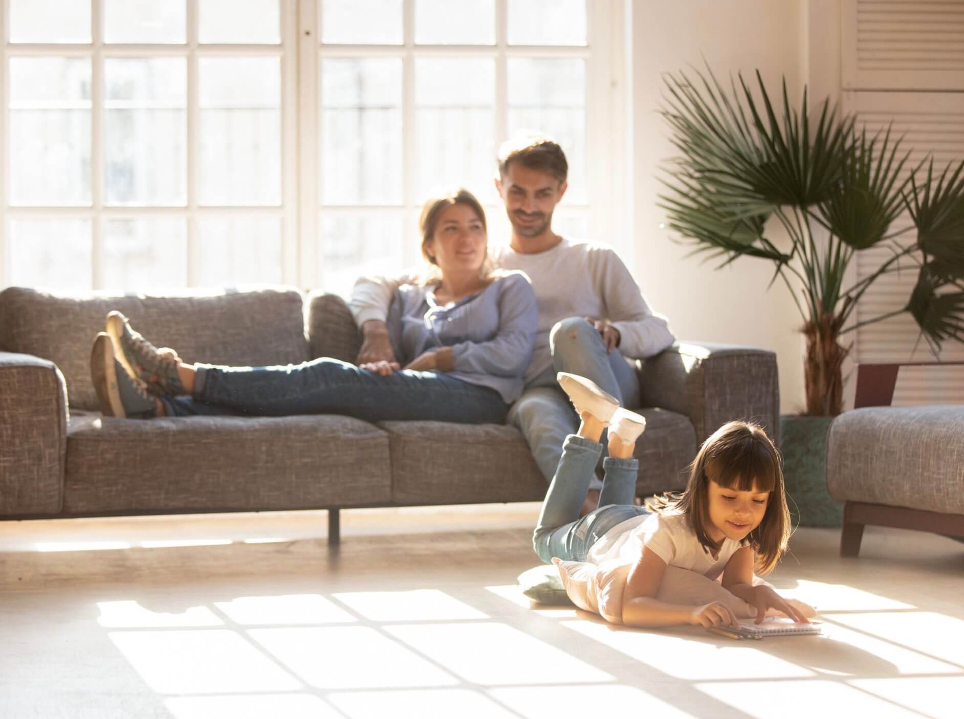 A family relaxing in a sun-drenched living room. The child lays on the floor reading a book while the parents watch happily from the couch.