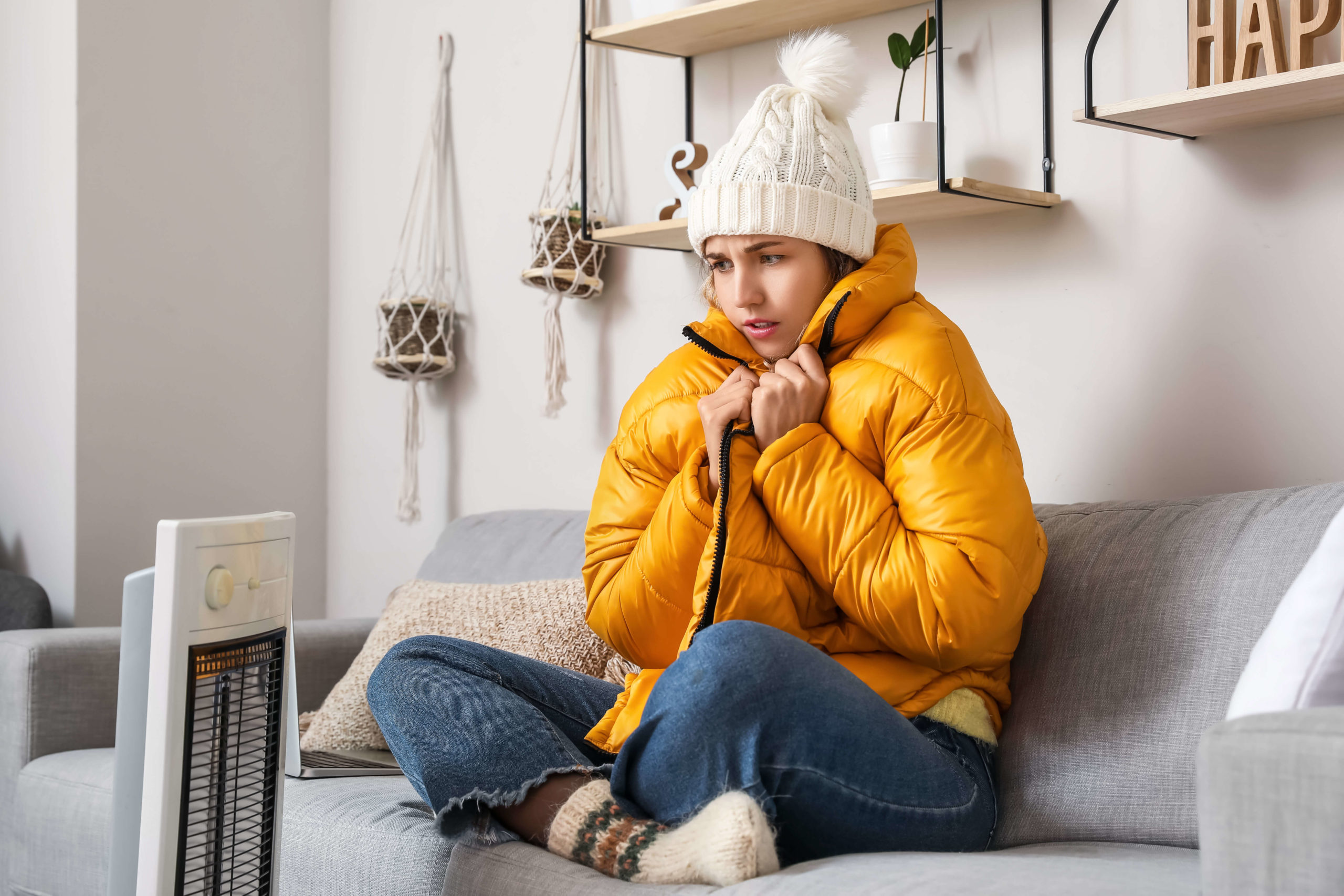 A person wearing a winter hat and coat inside the house, sitting in front of a space heater.