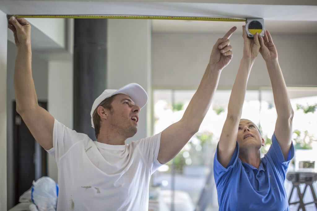 Couple, arms raised, using tape measure to measure ceiling