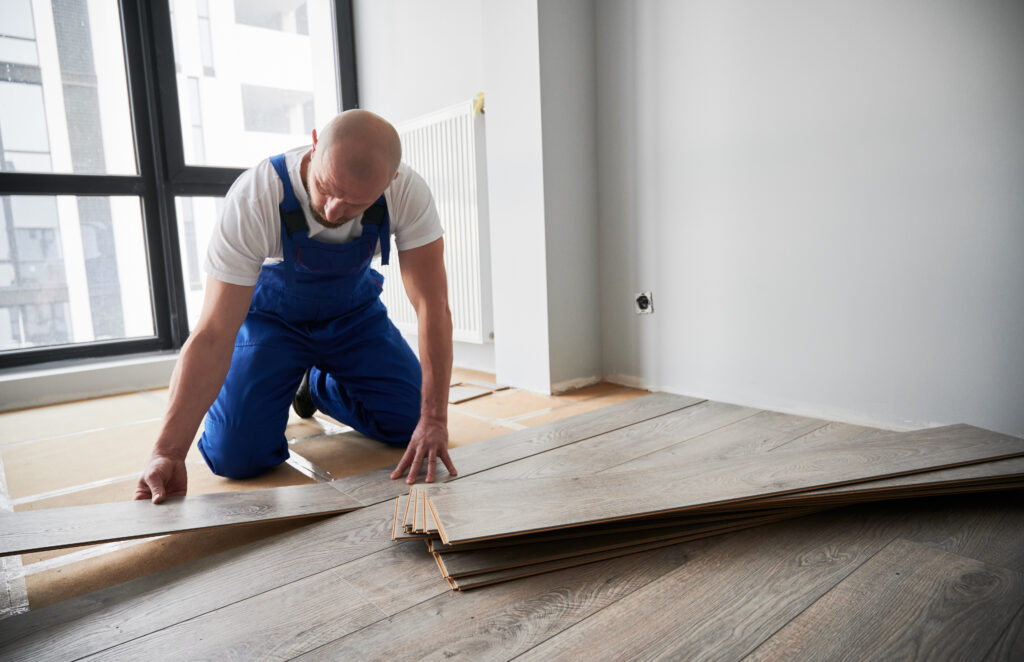 Man in work overalls installing laminate wood flooring in new flat. Male construction worker laying laminate wooden planks on floor underlayment. Home renovation concept.